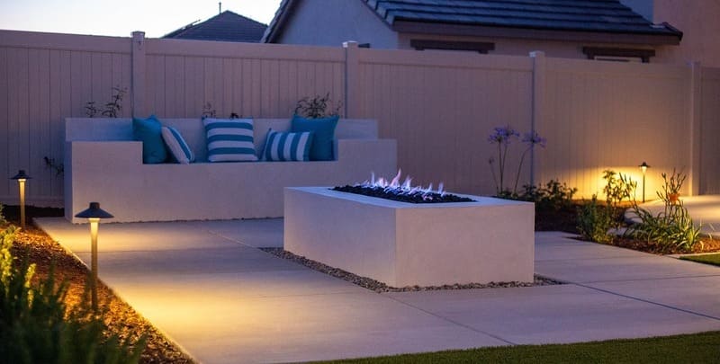 Backyard setting with outdoor lighting surrounding a concrete fire pit and a seat with blue and white striped cushions.